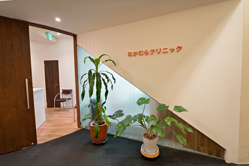 なかむらクリニックの入口（本町駅 徒歩3分・カネセ中央ビル1階）。クリーム色の壁に朱色のクリニック名サインと観葉植物